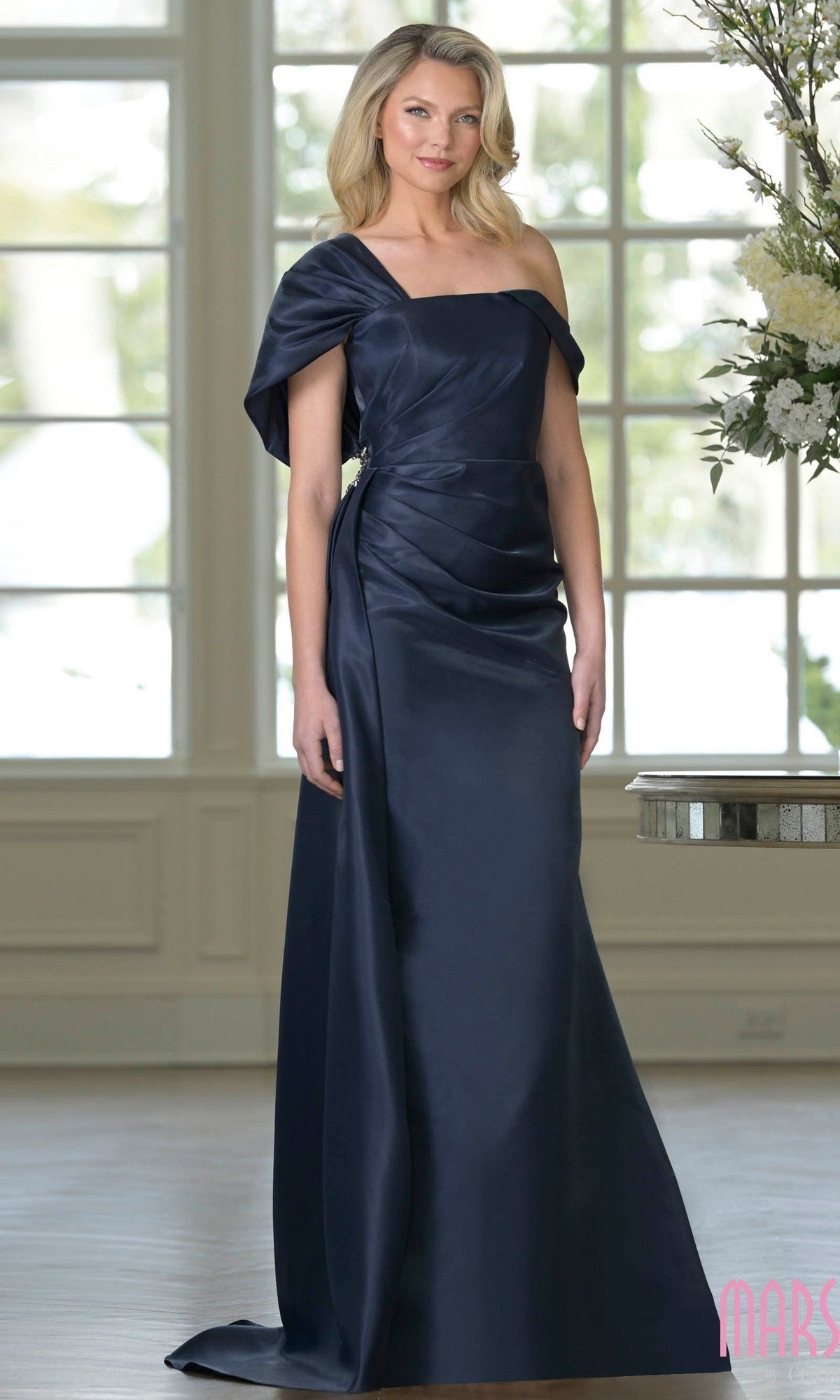 A woman wearing the Marsoni MV1405 long formal dress in Midnight stands indoors, framed by a window and floral arrangement in the background.